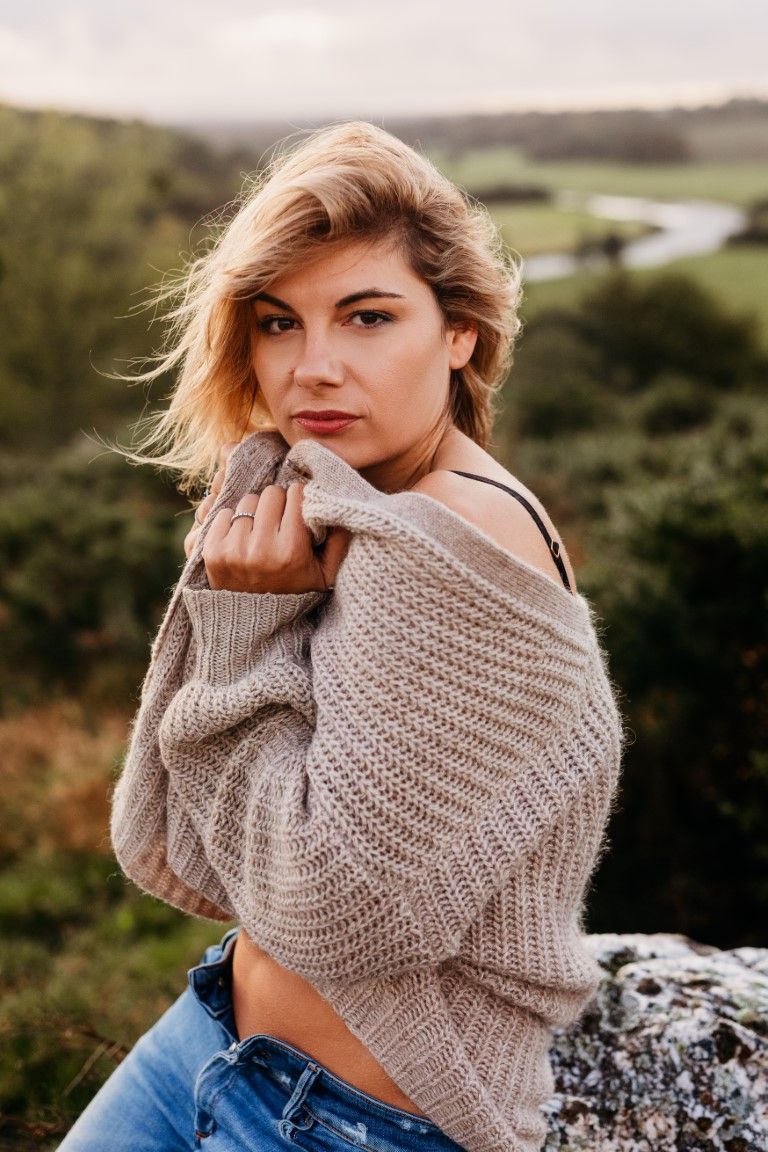 Young blonde woman in beige knit sweater and jeans poses outdoors with scenic green valley backdrop.