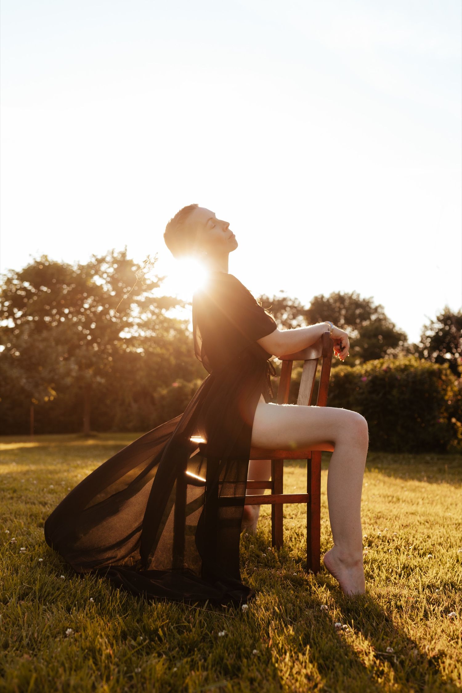 A woman in a flowing black dress sits on a wooden chair outdoors, bathed in golden sunset light.