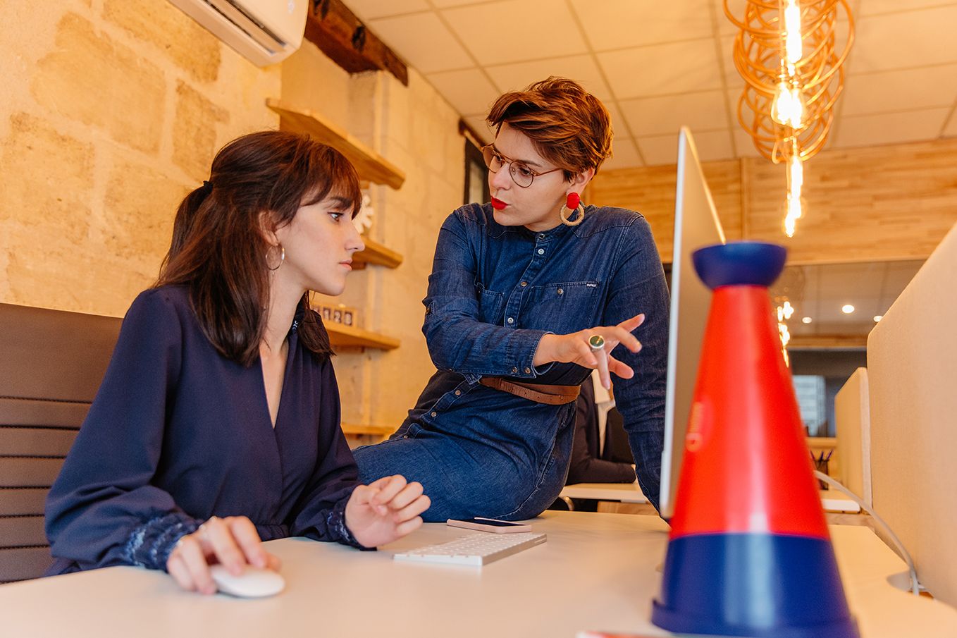 Two women collaborating at a desk with a computer, colorful cone decoration in the foreground.