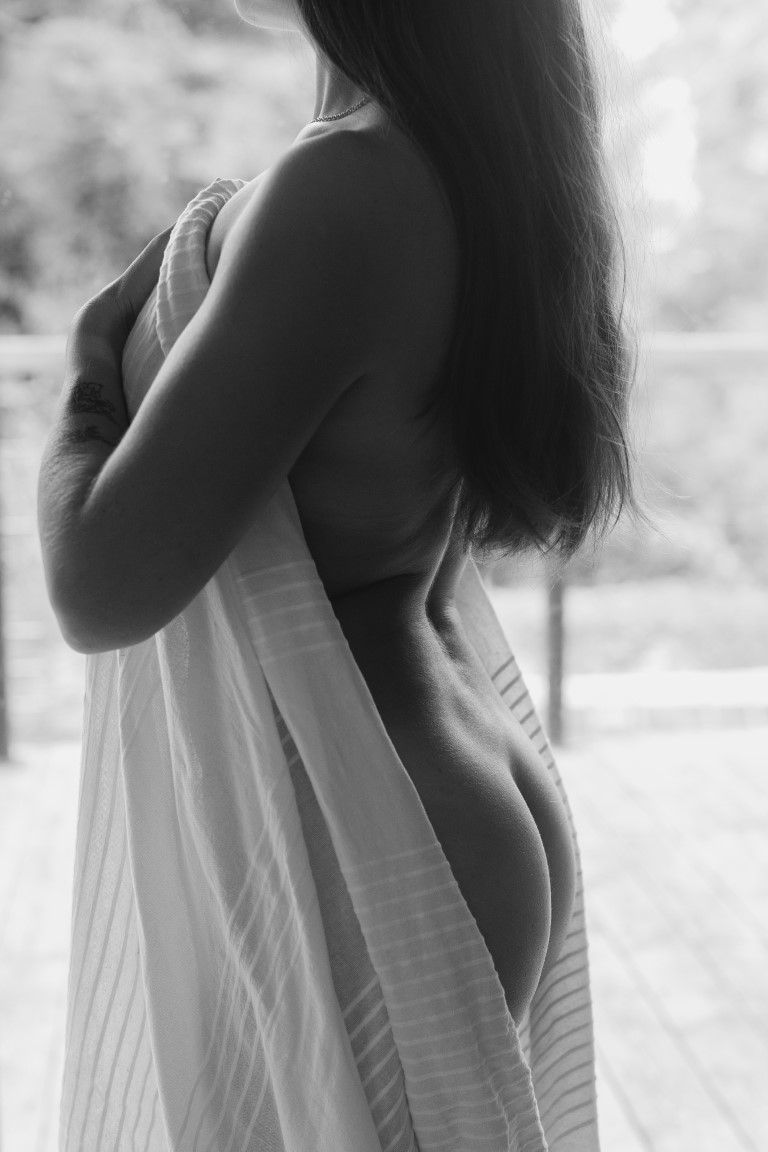 Black and white boudoir photo of a woman draped in a white striped sheet, showing her bare back.