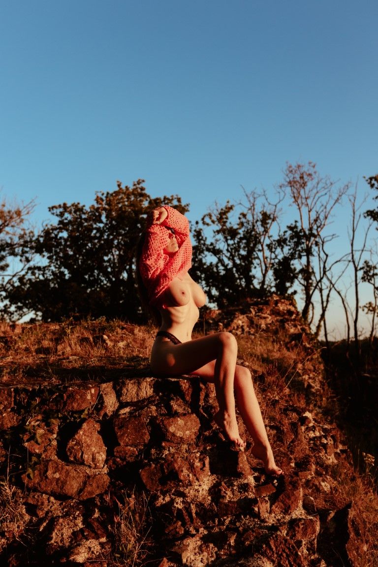 A nude woman with a red wrap on her head sits on rocky terrain at golden hour with trees and blue sky behind her.