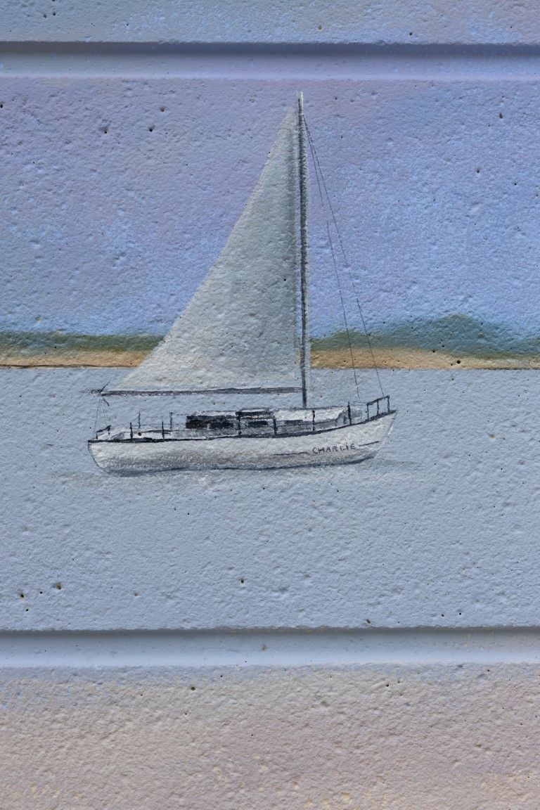 A painted mural of a sailboat on a light blue brick wall, depicting a serene nautical scene with calm waters and sandy shore.