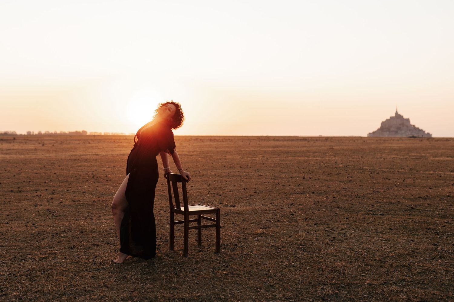 Woman leaning on a wooden chair in an open field at sunset with Mont Saint-Michel silhouette in background.