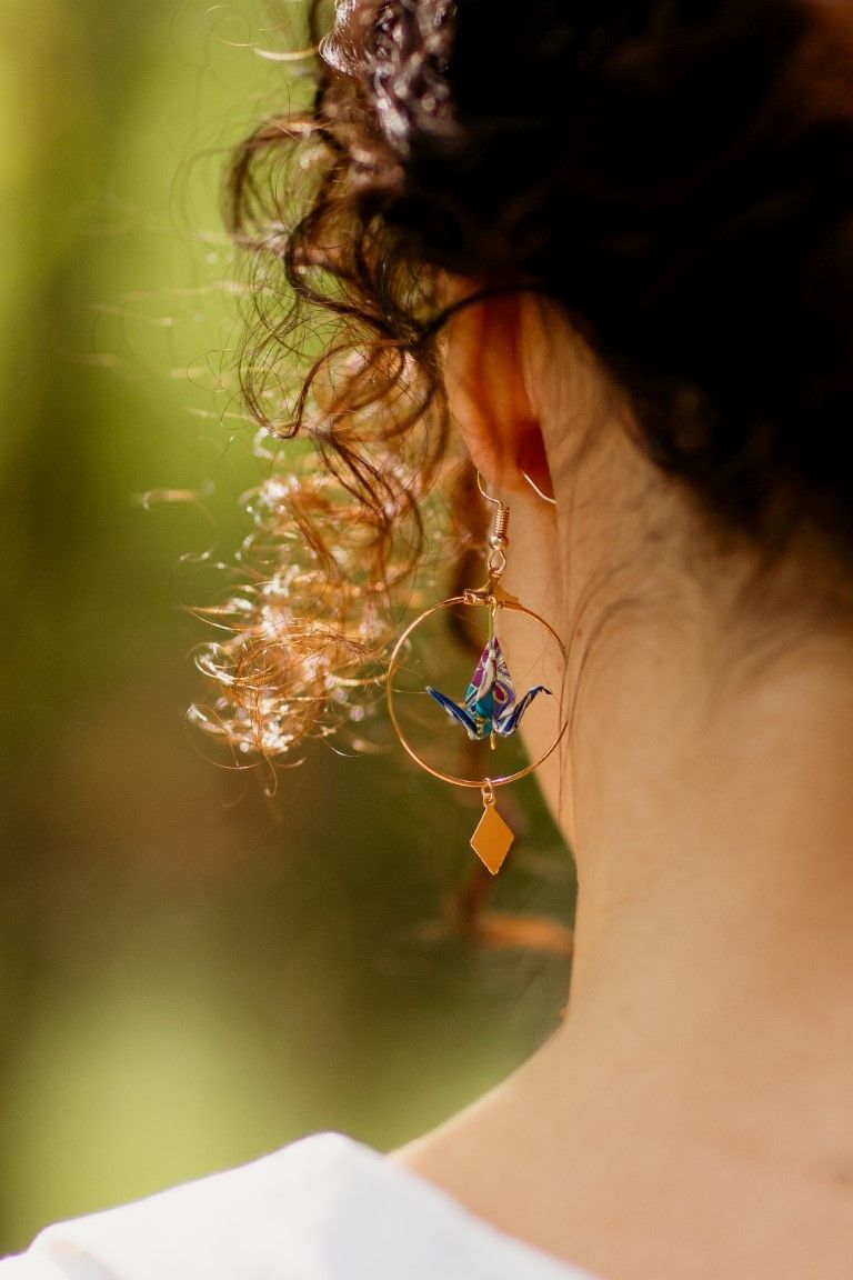 Close-up of gold hoop earring with blue origami crane pendant on a woman.