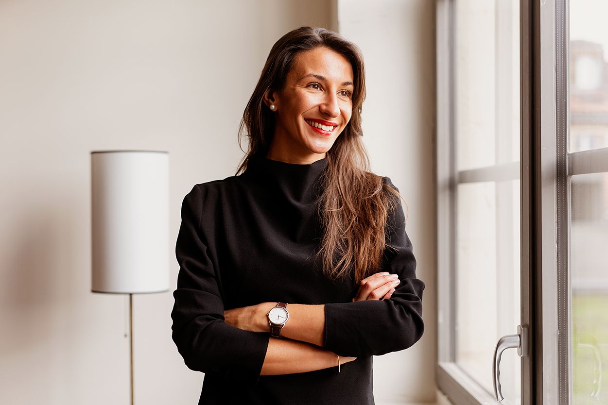 Brunette woman in black turtleneck standing with arms crossed, smiling near window indoors.