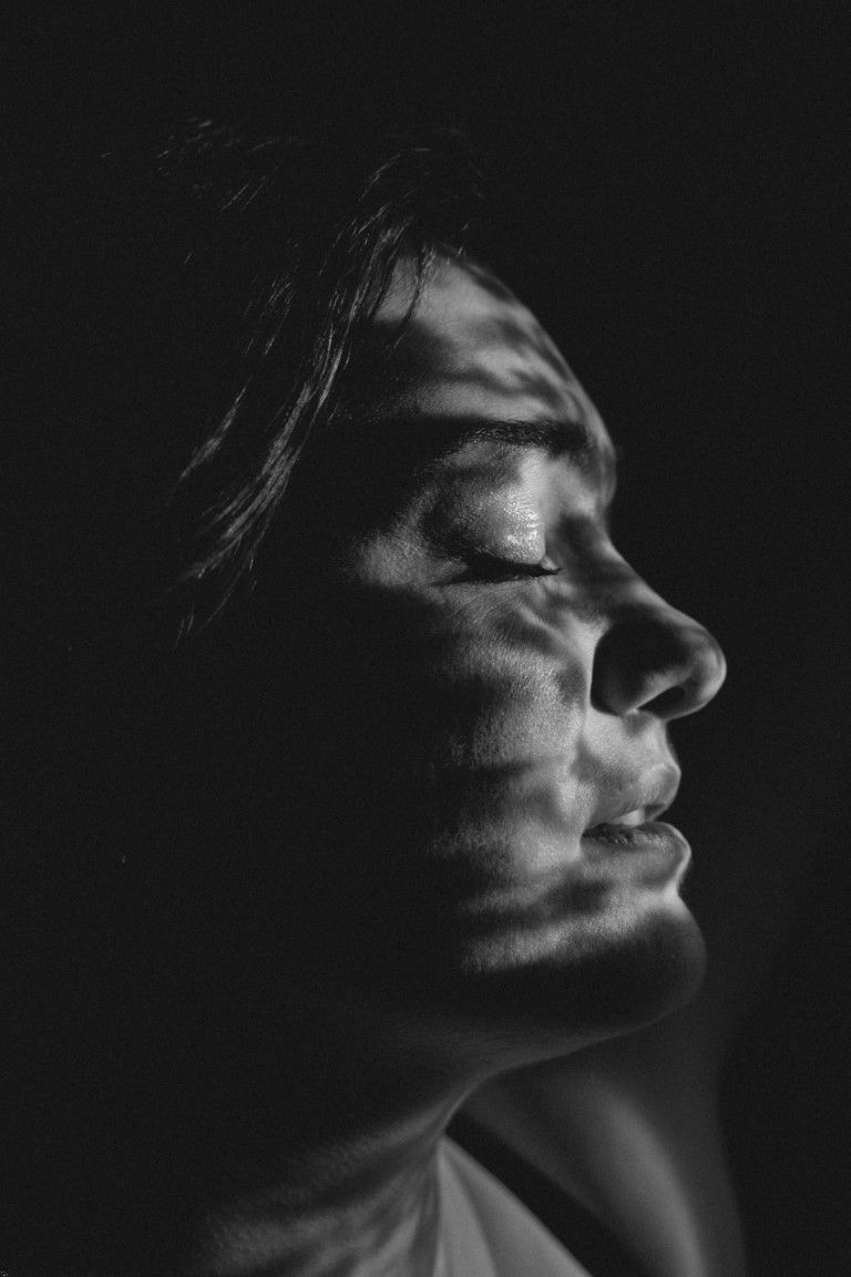 Dramatic black and white close-up portrait of woman with closed eyes and shadow patterns on face.