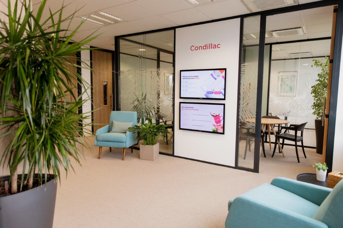 Modern Condillac office reception with teal chairs, green plants, and digital display screens.