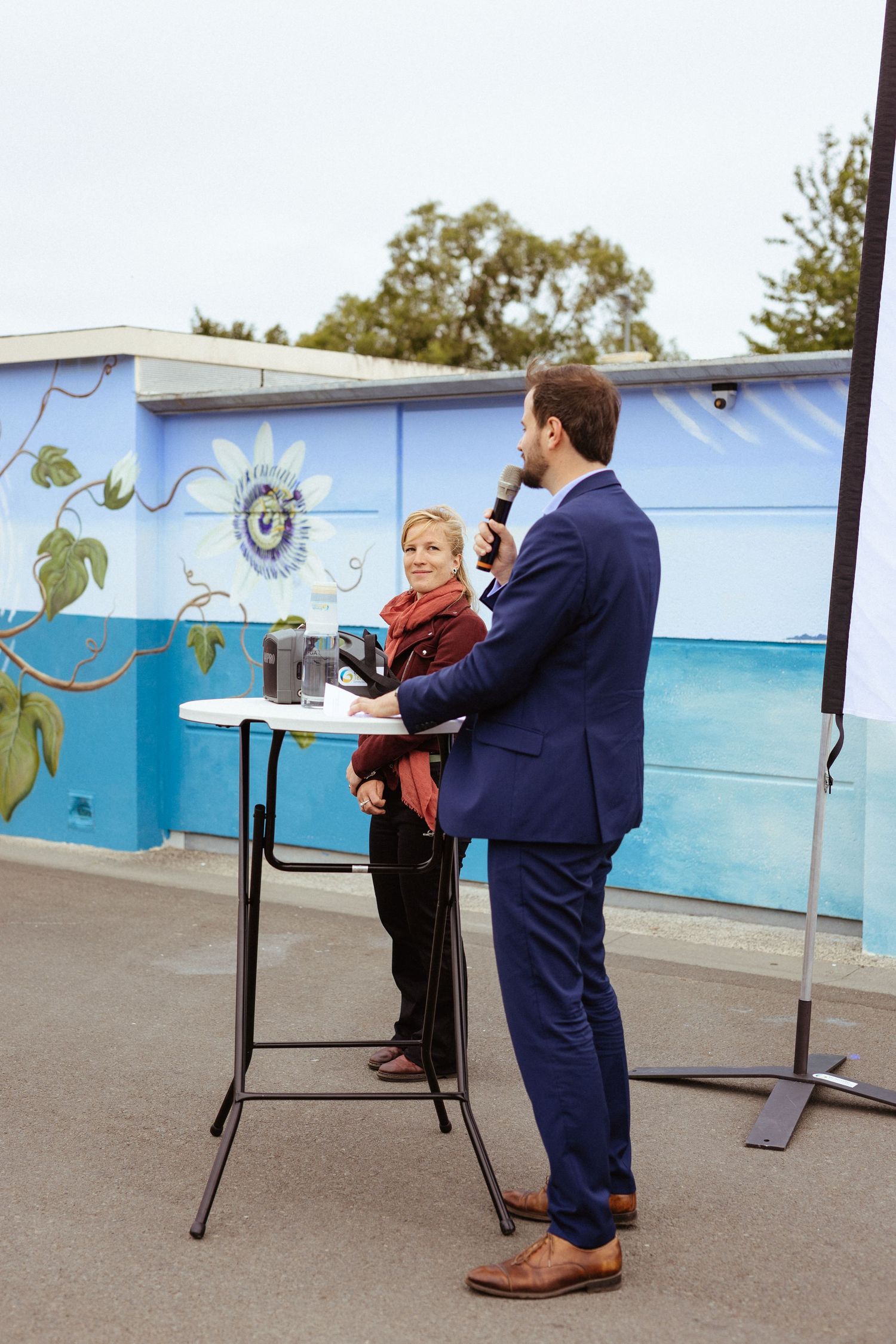 Man in blue suit speaking into microphone outdoors near a colorful mural, with a woman standing beside a high table.