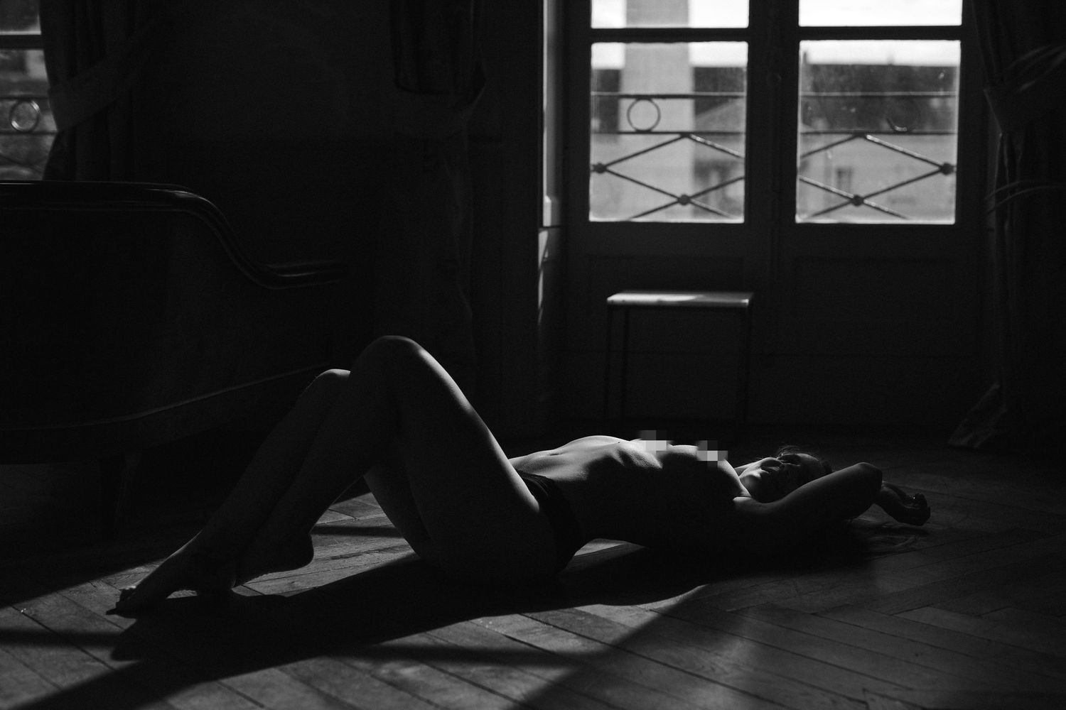 Black and white artistic nude photo of a person lying on a wooden floor near sunlit windows.