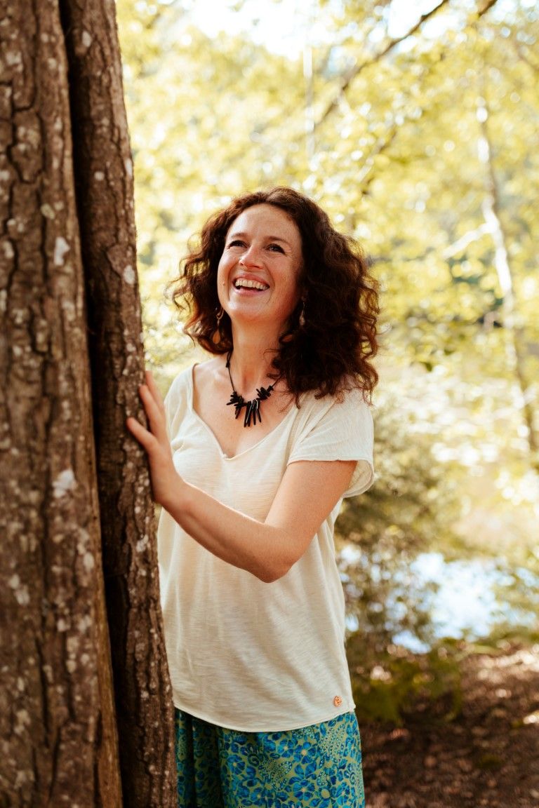 Smiling woman with curly hair leaning against a tree trunk in a sunlit forest, wearing a white top and floral skirt.