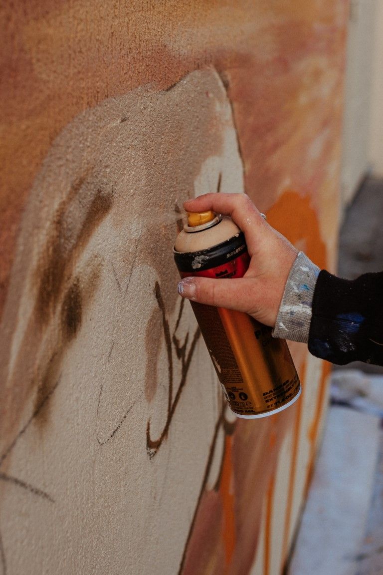 A hand holding a gold spray paint can creating graffiti art on a textured wall with orange and beige tones.