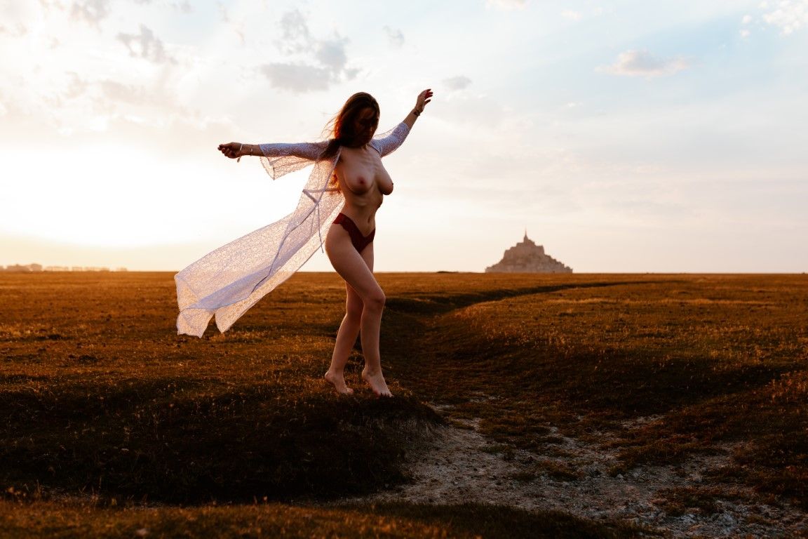Woman in open white robe posing at sunset with Mont Saint-Michel castle in the background.