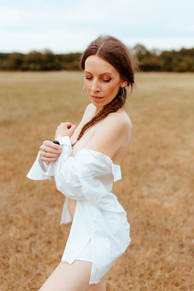 Woman in white off-shoulder shirt poses gracefully in golden field with trees in background.