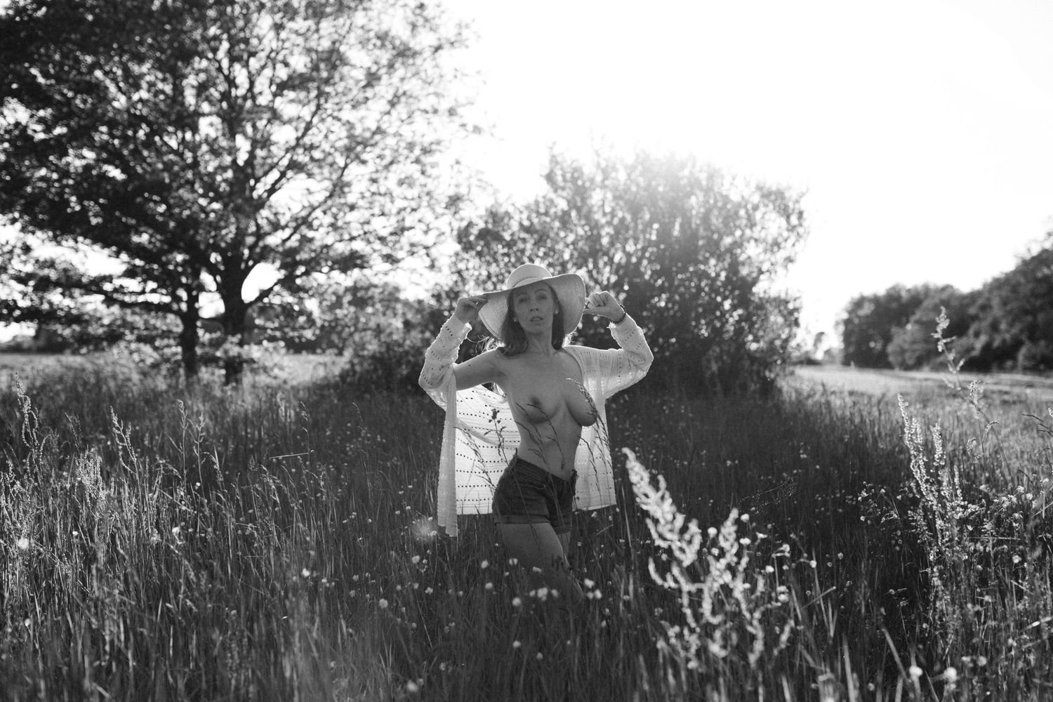 Black and white photo of a woman in a sun hat standing in a wildflower meadow with trees in the background.
