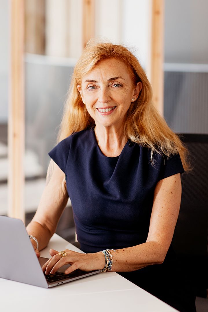 Mature businesswoman with red hair smiling while working on laptop at modern office desk.
