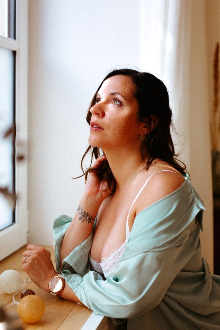 Brunette woman in lace bra and mint shirt gazing upward while sitting by a window.
