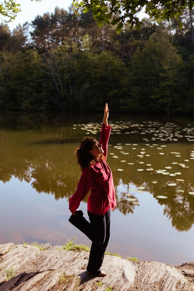 Woman in red shirt practicing tree pose yoga on rocks beside a serene lily pad pond surrounded by lush forest.