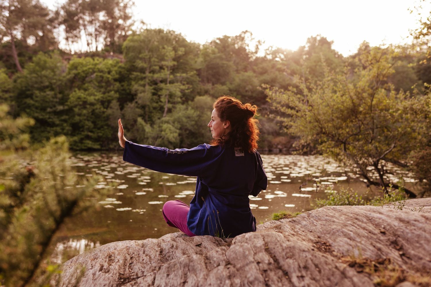 Woman practicing tai chi on a rock by a tranquil pond surrounded by lush green trees in nature.