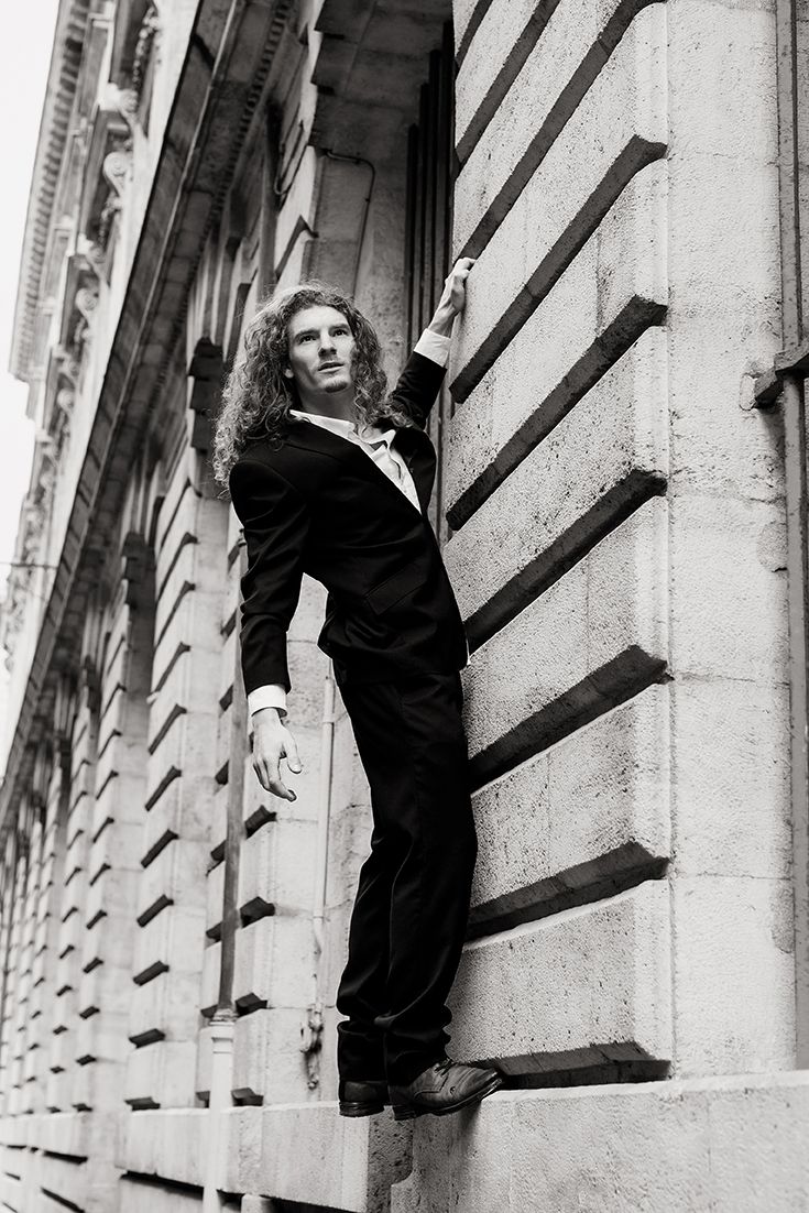 Élégante photo en noir et blanc d'un homme aux cheveux longs, vêtu d'un costume noir, appuyé contre la façade en pierre d'un bâtiment.