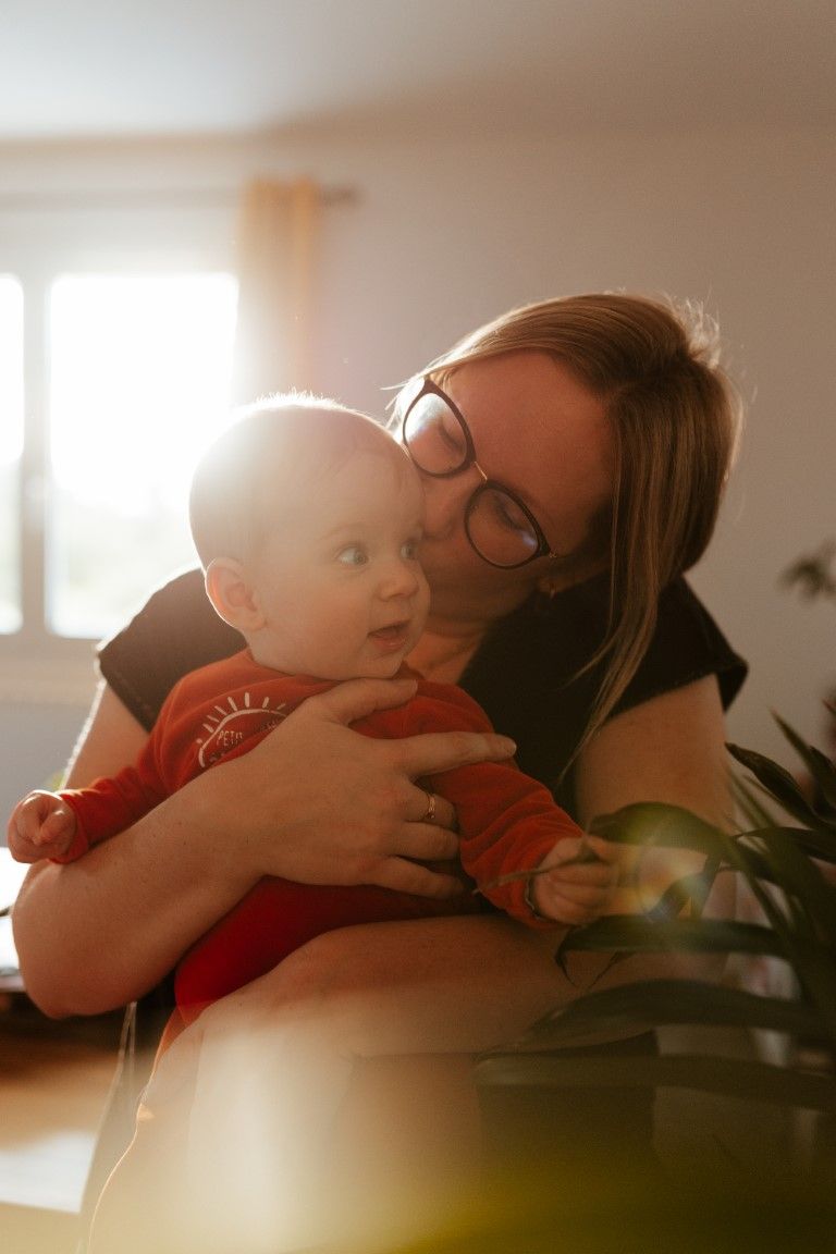 Une mère portant des lunettes embrasse son bébé souriant dans la douce lumière tamisée d'une pièce éclairée par le contre-jour.