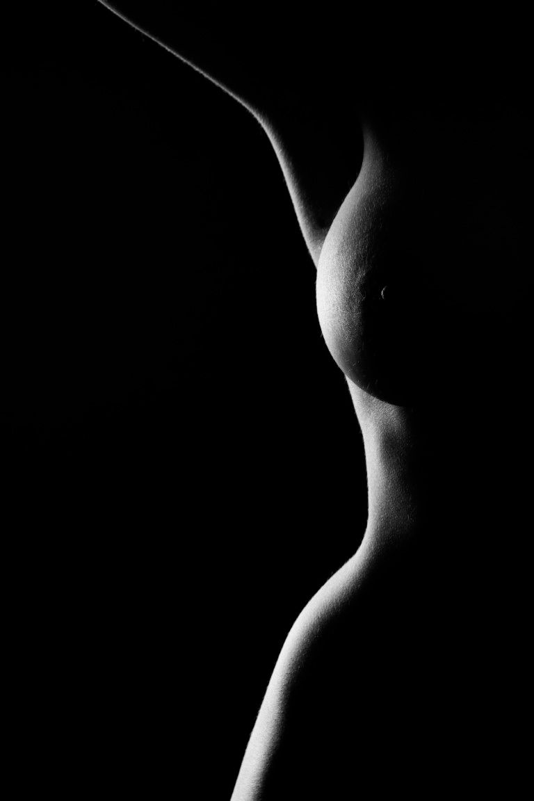 Black and white fine art nude side profile silhouette of a womans torso against dark background.