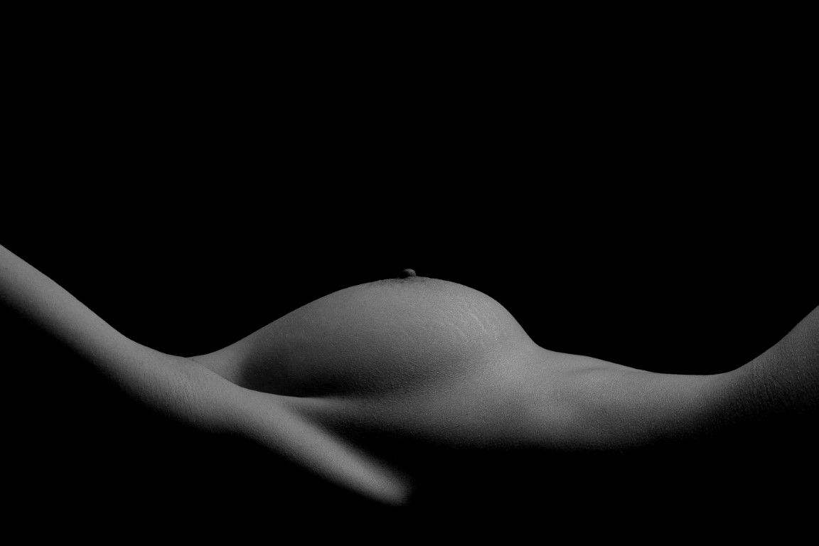 Black and white artistic nude photograph highlighting body curves with dramatic low-key lighting against dark background.