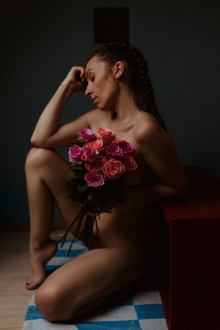 Woman sitting on floor holding bouquet of pink and orange roses in a moody, dimly lit studio setting.