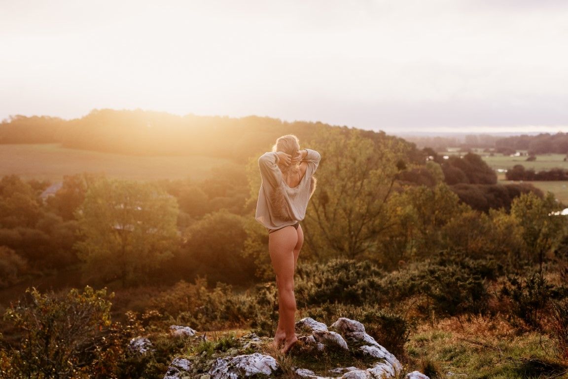 Person standing on rocky hilltop at golden sunset overlooking lush green valley and distant coastline.