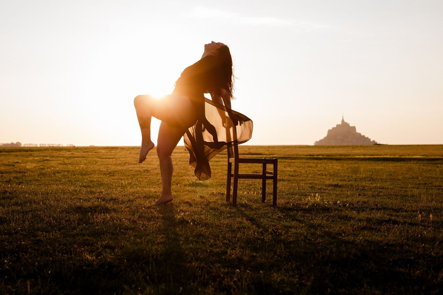 Silhouette of a woman posing by a chair at golden sunset with Mont Saint-Michel in the background.