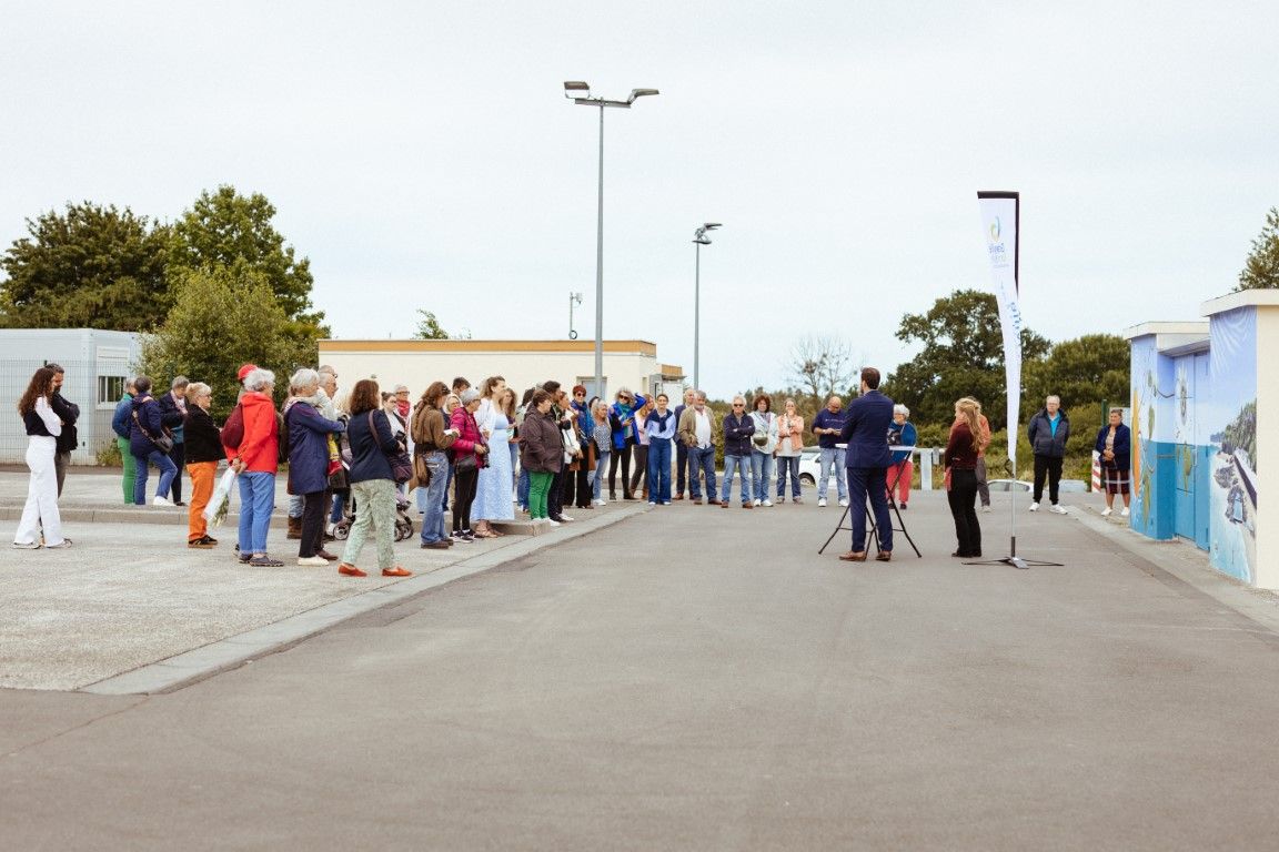 Large group of people gathered outdoors near a coastal facility for a community event on an overcast day.