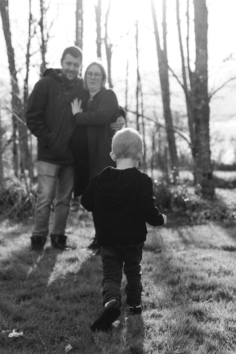 Image en noir et blanc d'un jeune enfant marchant vers ses parents souriants, debout parmi les arbres d'un parc.