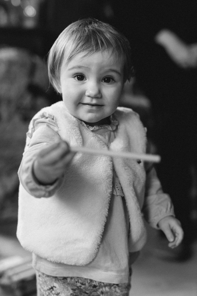 Une adorable petite fille en gilet duveteux tend un bâton vers l'objectif, en noir et blanc.