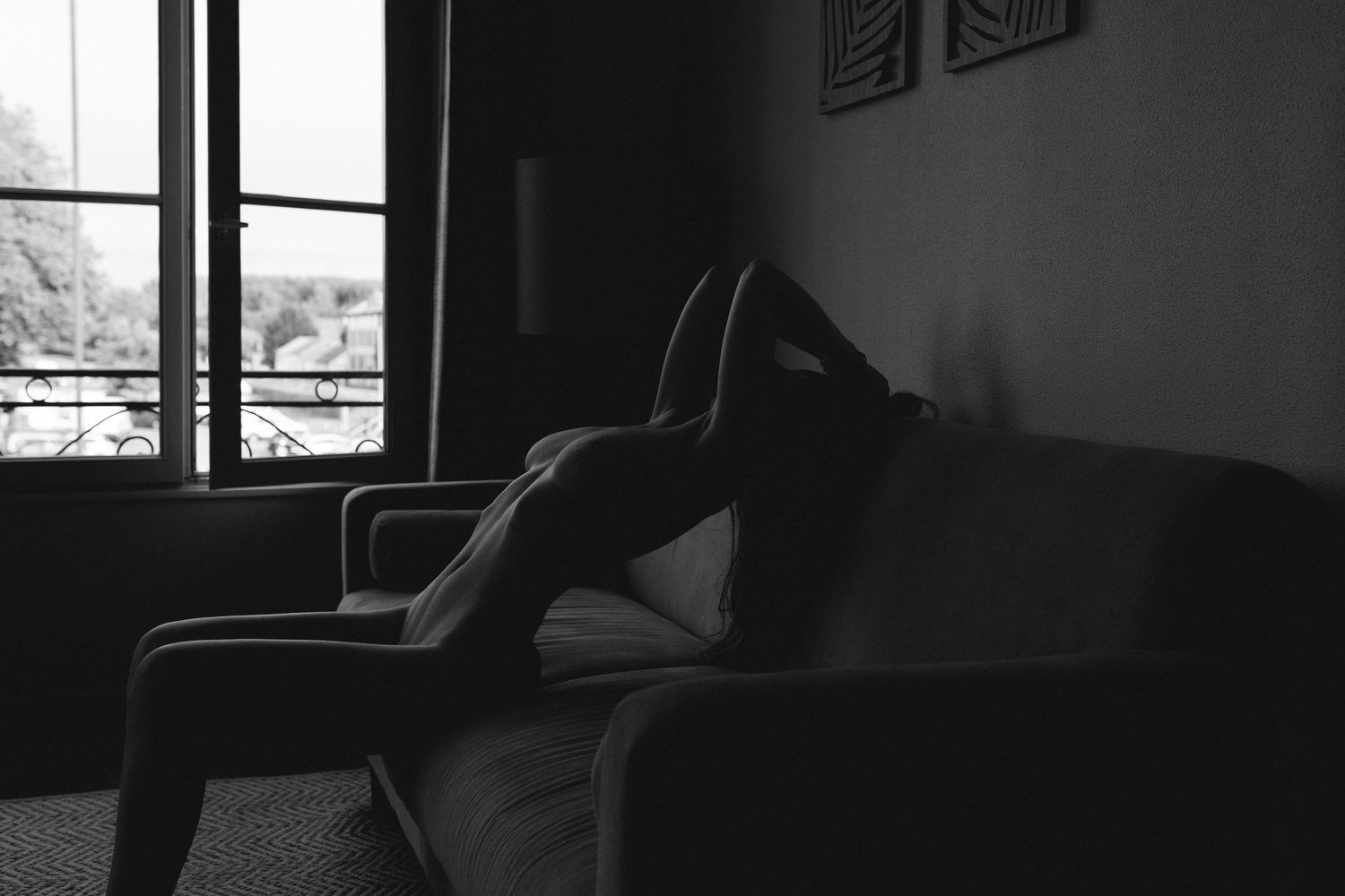 Black and white photo of a person sitting alone on a sofa in a dimly lit room near a window.