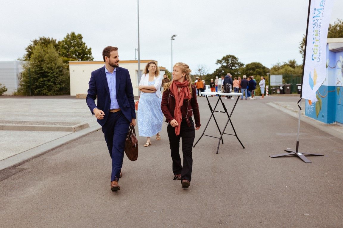 A man in a blue suit and woman in burgundy jacket walk together outdoors at an event venue on a sunny day.