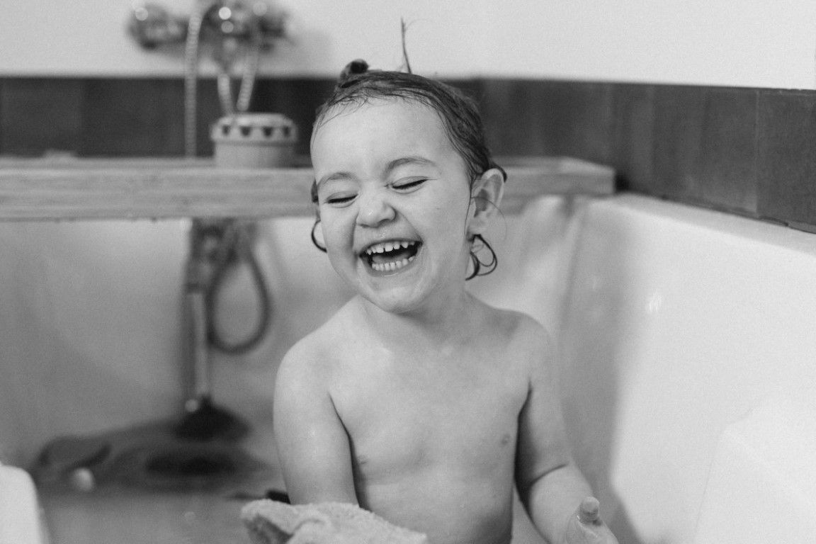 Petit enfant joyeux riant aux éclats, les yeux fermés, assis dans une baignoire, en noir et blanc.