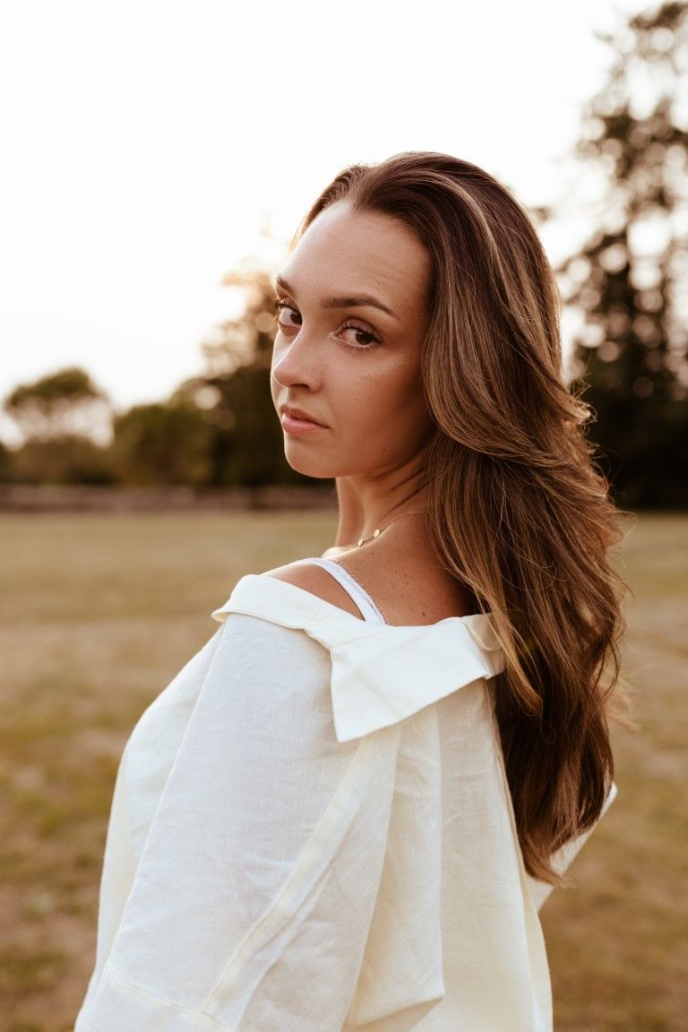 Jeune femme brune en haut blanc à épaules dénudées posant à l'extérieur sous la lumière dorée du crépuscule, avec des arbres en arrière-plan