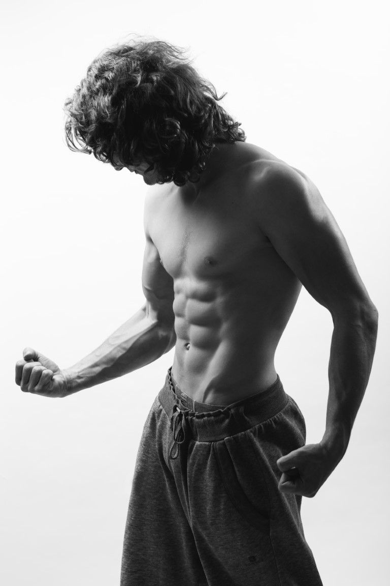 Photo en noir et blanc d'un homme musclé, torse nu, aux cheveux bouclés, faisant des flexions en pantalon de survêtement gris.