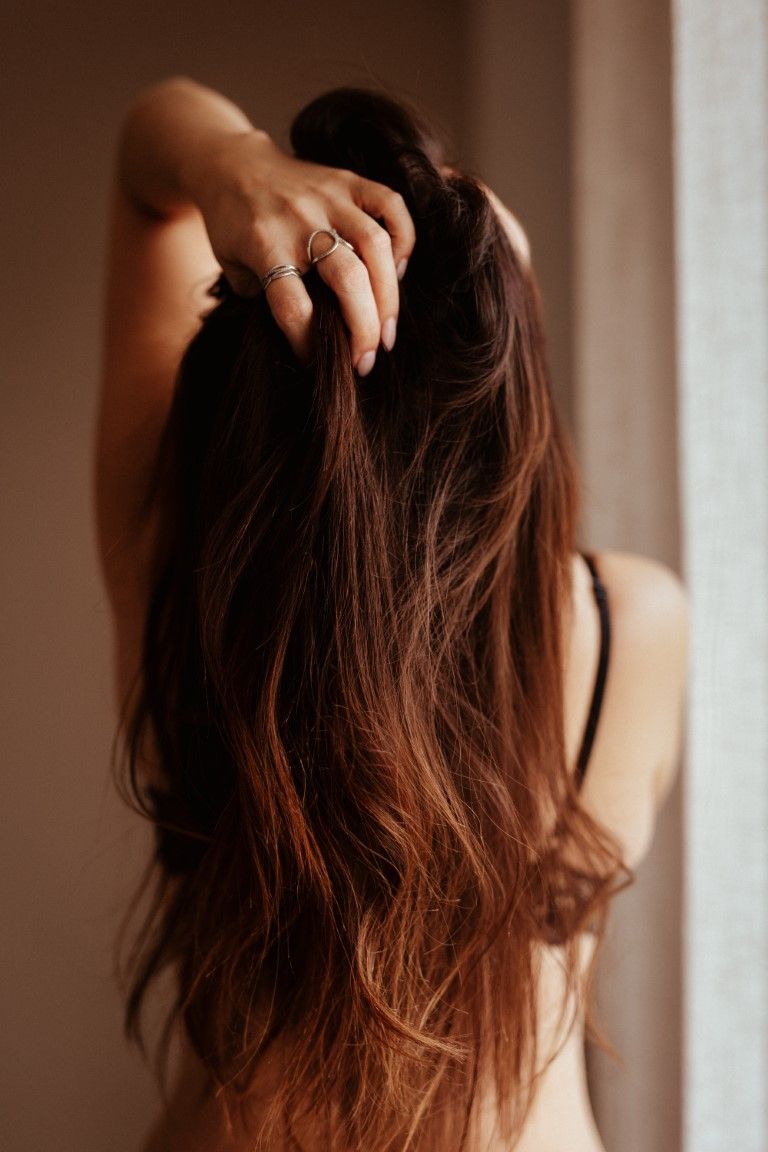Back view of a woman with long brunette hair holding it up with ringed fingers near a window.