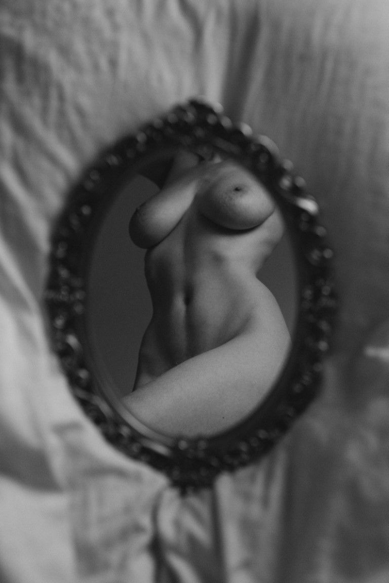Corps nu en noir et blanc se reflétant dans un miroir ovale orné sur fond de tissu blanc.