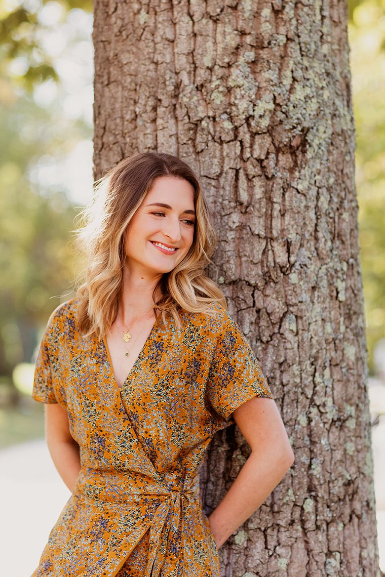 Smiling woman in yellow floral wrap dress leaning against tree trunk in sunny park.