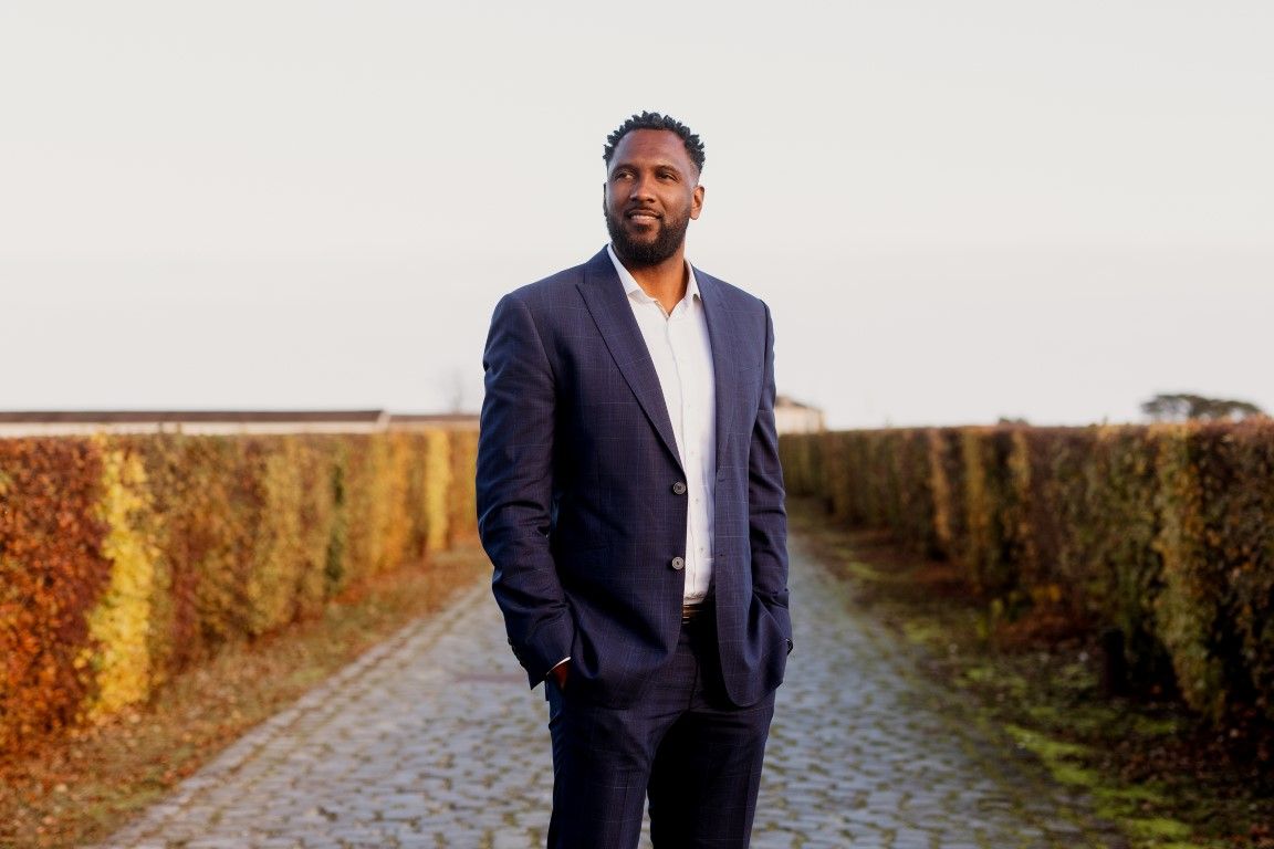 Tall man in navy suit standing on cobblestone path between autumn hedgerows outdoors.