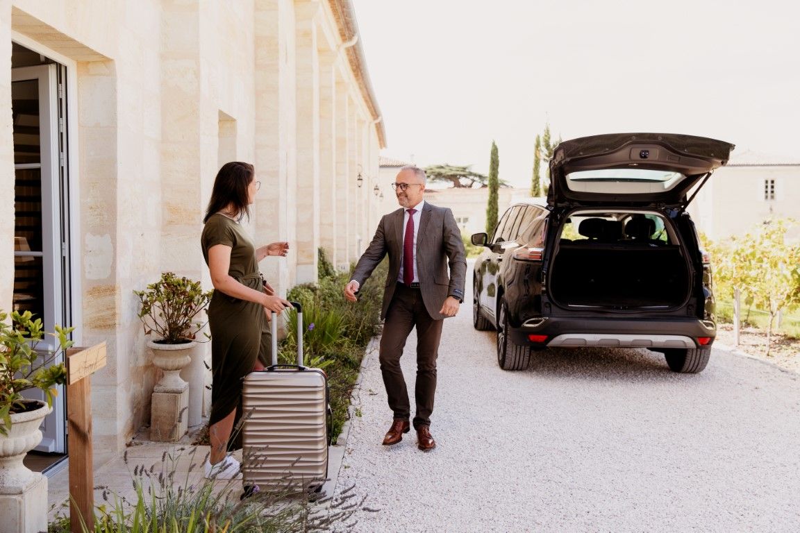 Chauffeur greeting female passenger with luggage outside stone building near black SUV.