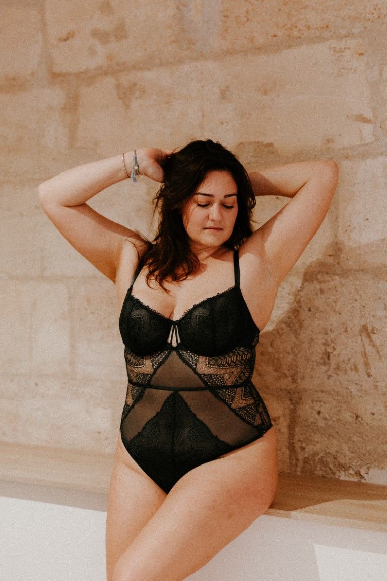 A curvy woman poses confidently in a black lace bodysuit against a stone wall.