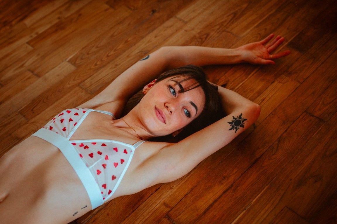 Young woman with star tattoo lying on hardwood floor wearing white heart-print lingerie.