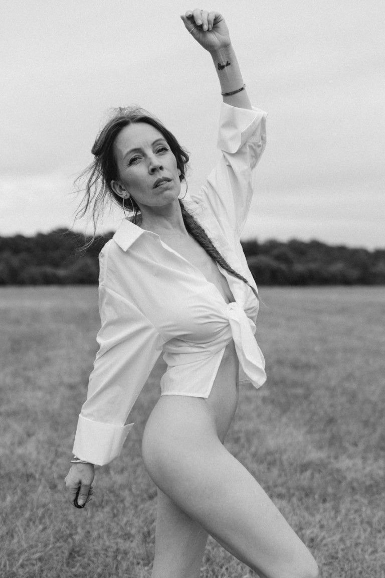 Photo en noir et blanc d'une femme en chemise blanche nouée posant de manière dynamique dans un champ ouvert.