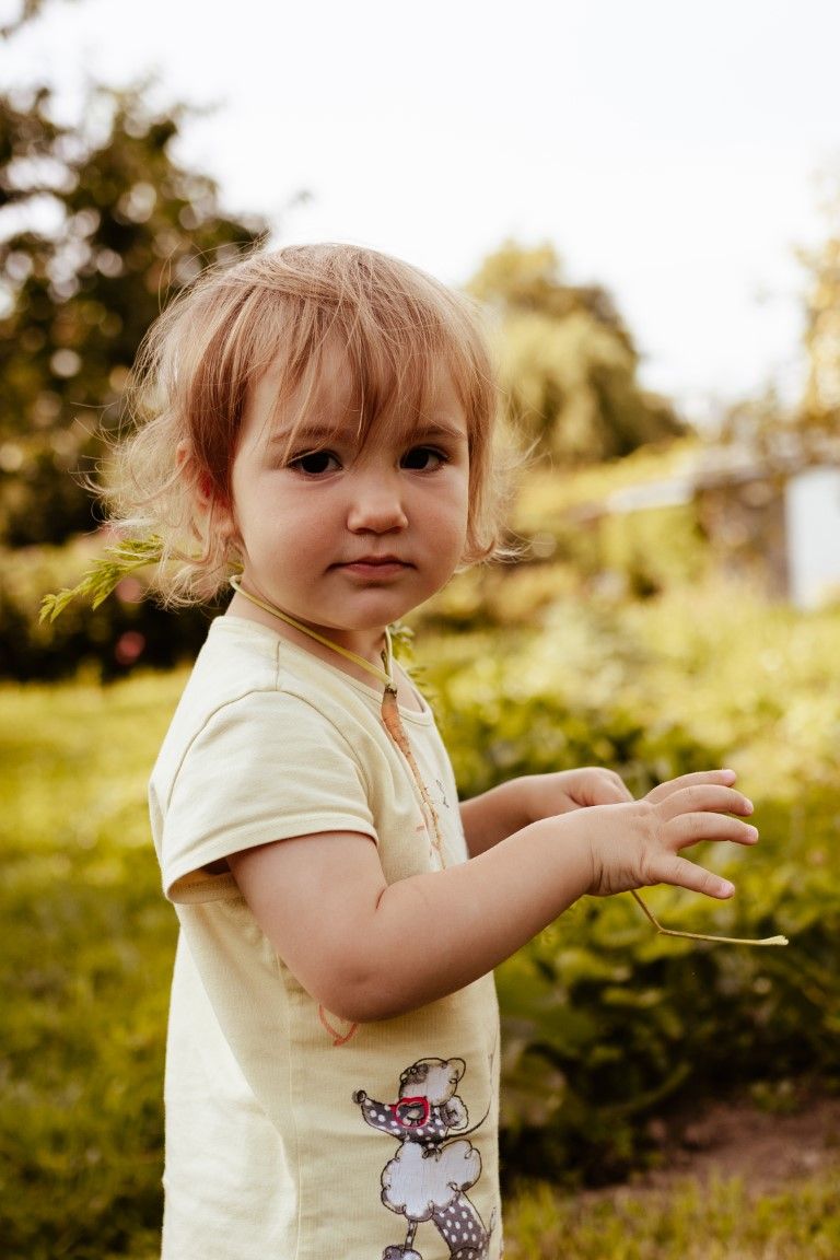 Une adorable petite fille blonde en t-shirt blanc se tient debout dans un jardin verdoyant et ensoleillé, tenant une petite brindille.