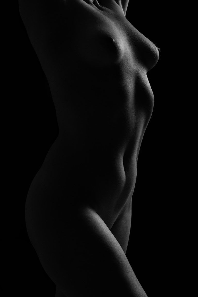 Black and white fine art nude photography highlighting the elegant curves of a female torso against dark background.