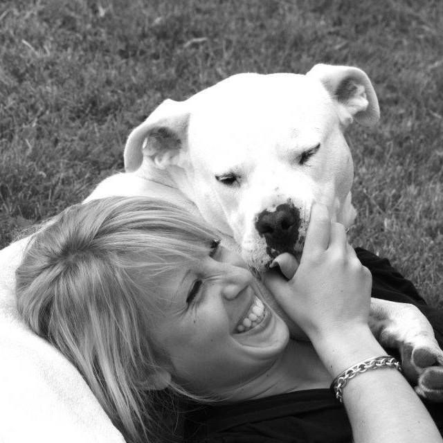 Une femme blonde rit tandis qu'un boxer blanc lui lèche le visage alors qu'elle est allongée dans l'herbe.