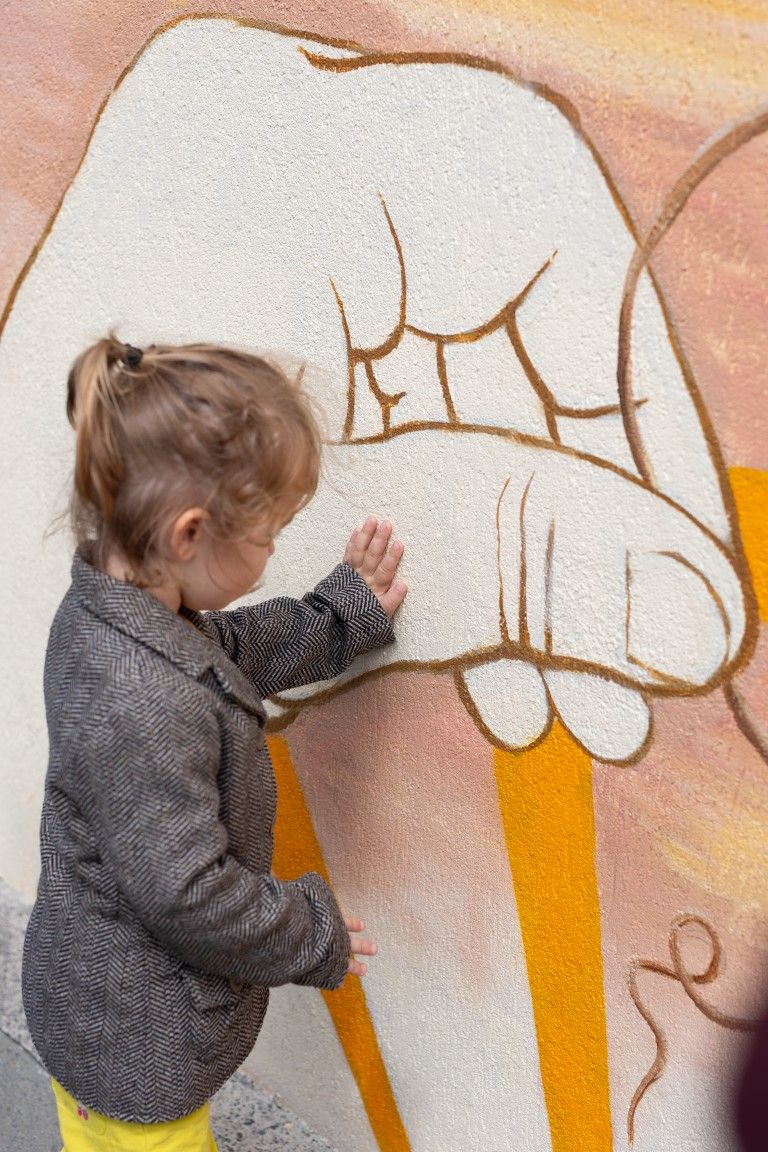 A toddler in a gray jacket touches a colorful graffiti mural featuring large painted hands on a textured wall.
