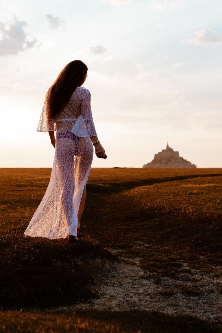 Femme en robe de dentelle blanche, debout sur un rocher, contemplant le Mont Saint-Michel à l'heure dorée.
